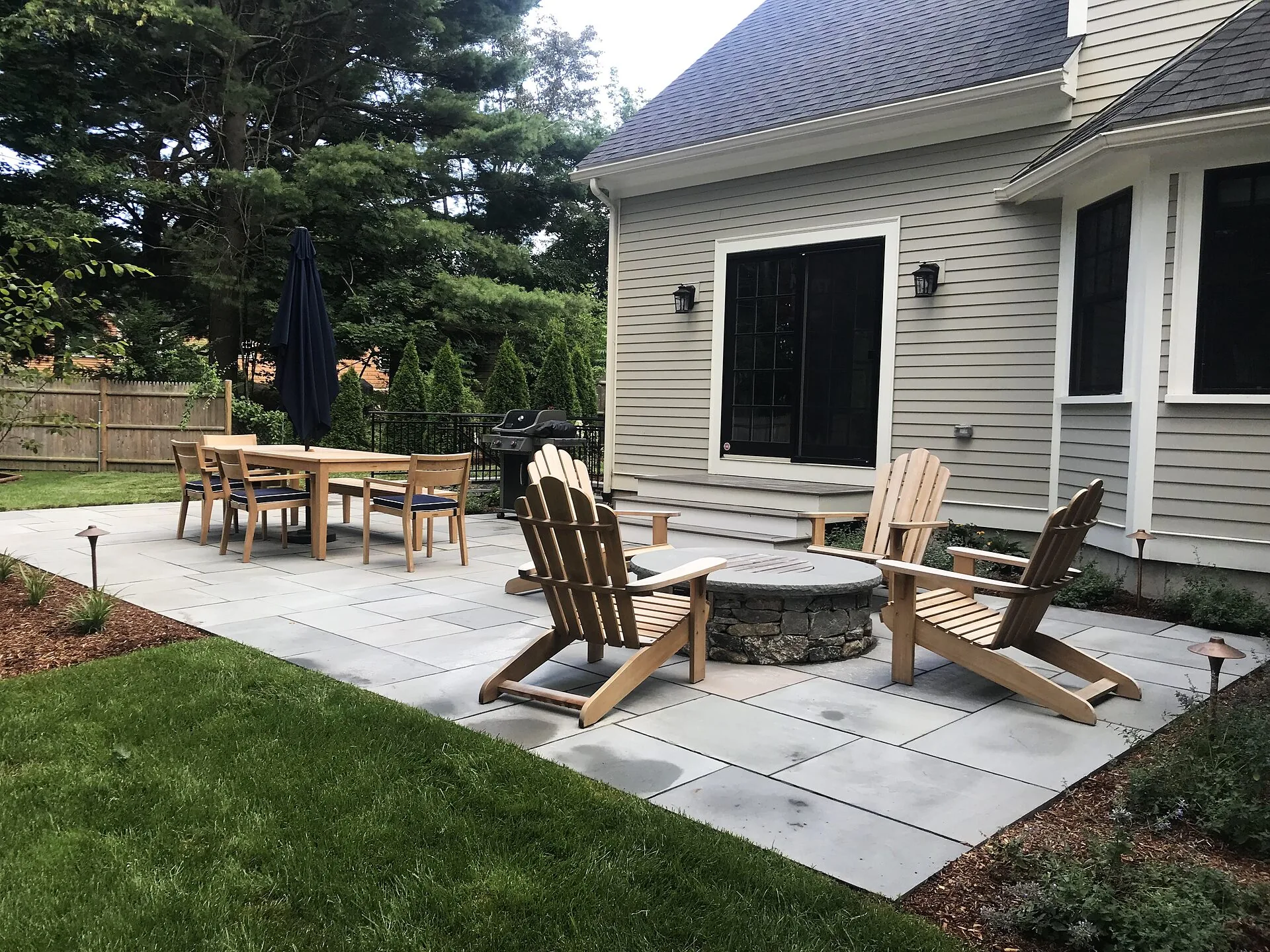 Bluestone patio with stone fire pit, wooden Adirondack chairs and a dining set beside a gray clapboard house