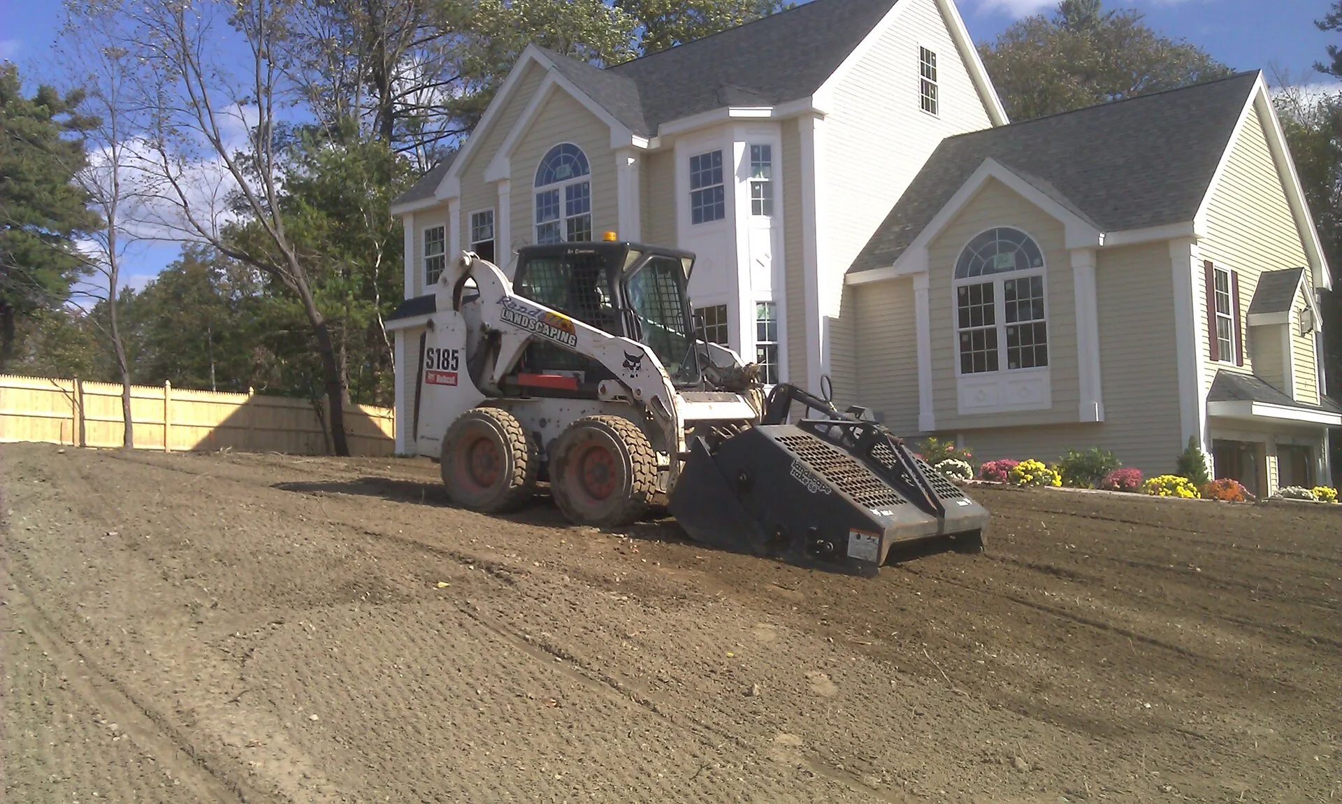 Bobcat S185 skid steer with a soil conditioner attachment grading bare dirt in front of a large house