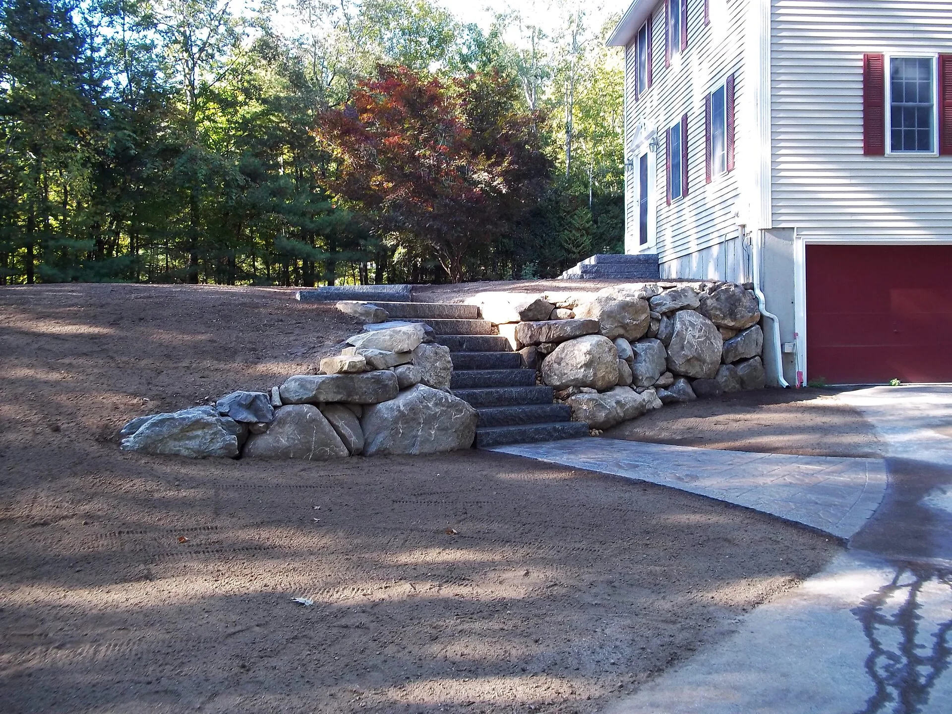 Boulder retaining wall with granite steps beside a house, leading up from a stamped concrete driveway and bare soil yard