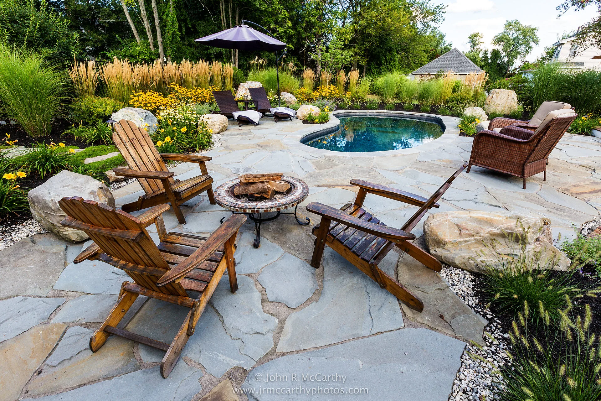 Flagstone patio with wooden Adirondack chairs around a fire pit, beside a small pool and ornamental grasses
