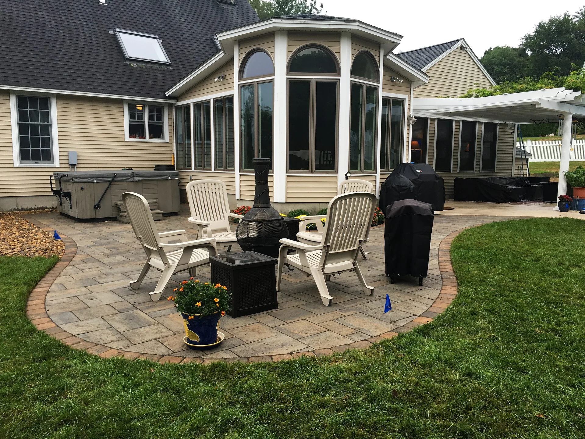 Curved paver patio with chairs, chiminea, grill and hot tub behind a house with sunroom and white pergola