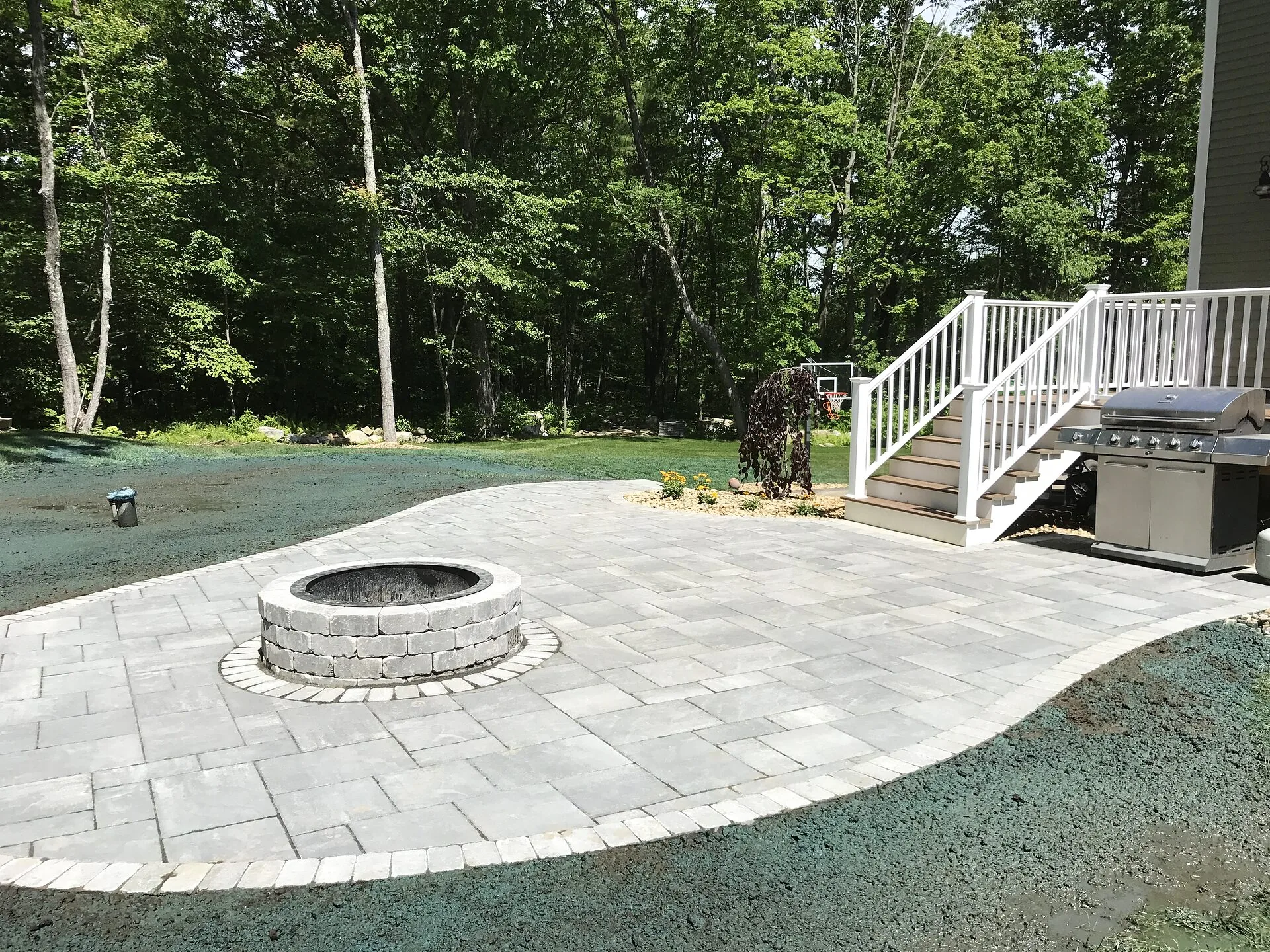 Curved gray paver patio with a round stone fire pit, next to white deck stairs and a stainless gas grill