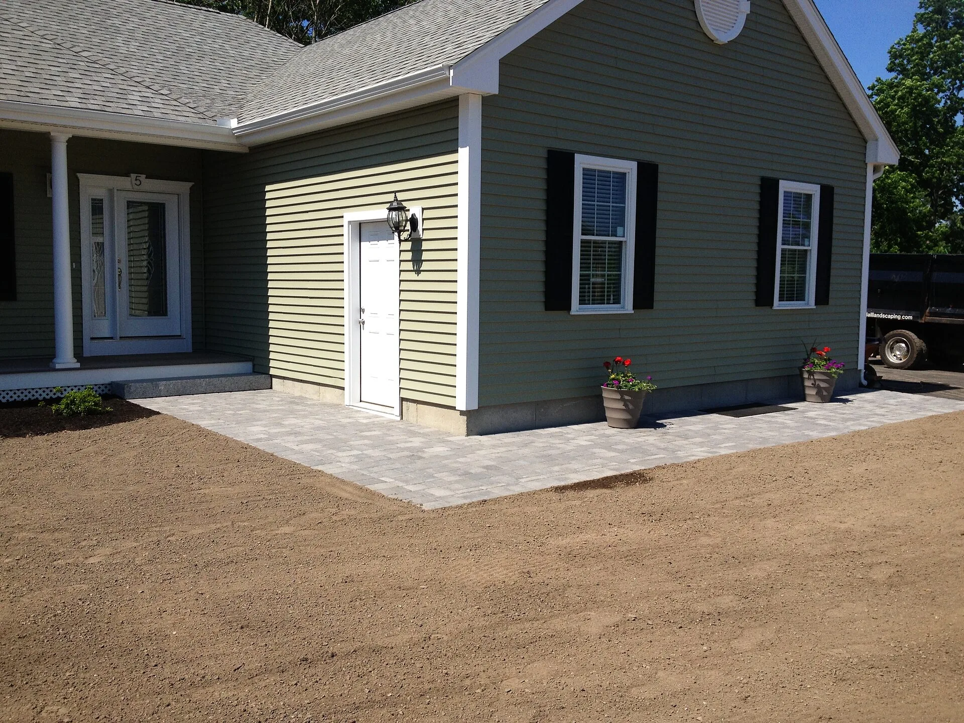 New paver walkway along a green sided house with potted red flowers and bare graded soil in the yard
