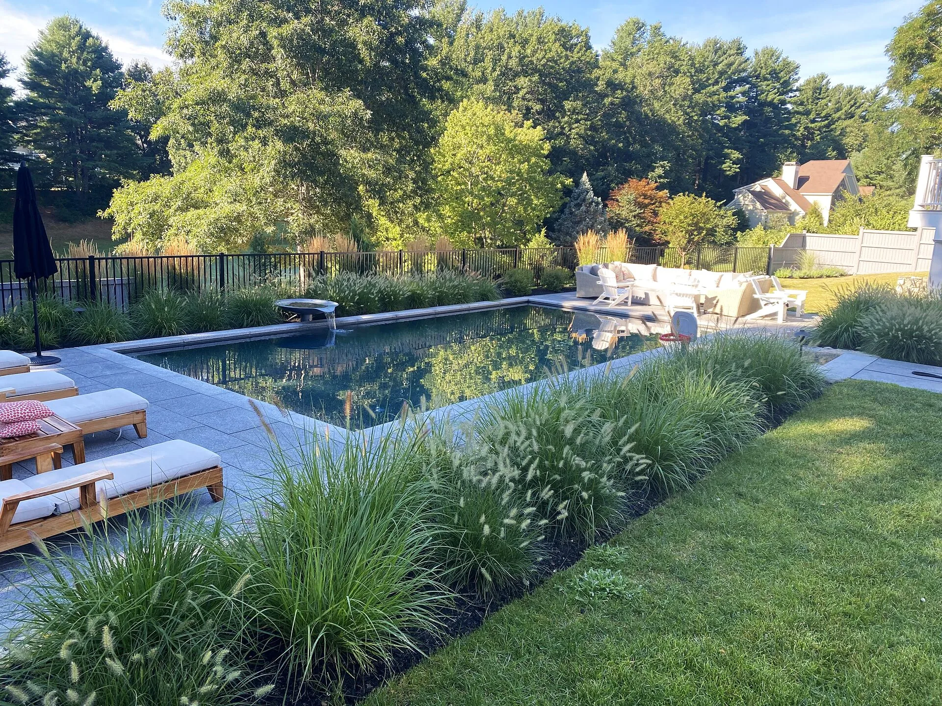 Backyard pool with stone deck, wooden loungers, patio seating and beds of ornamental grasses beside a lawn