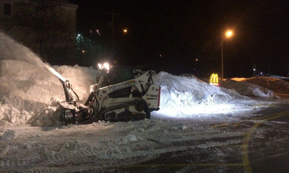 Bobcat track loader with snow blower attachment piling snow in a parking lot at night