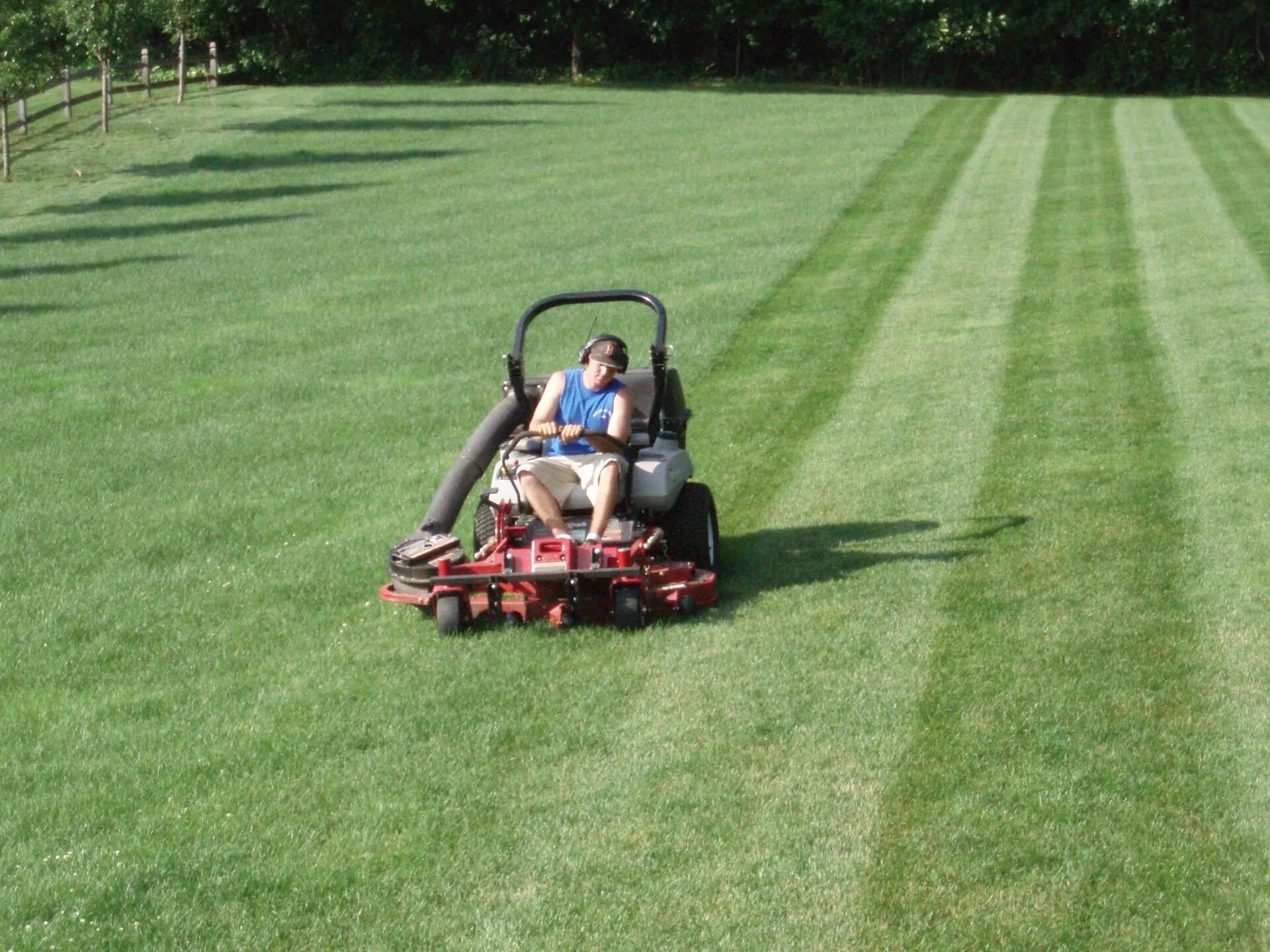 Man wearing ear protection drives a red zero turn riding mower across a large lawn with striped mowing lines